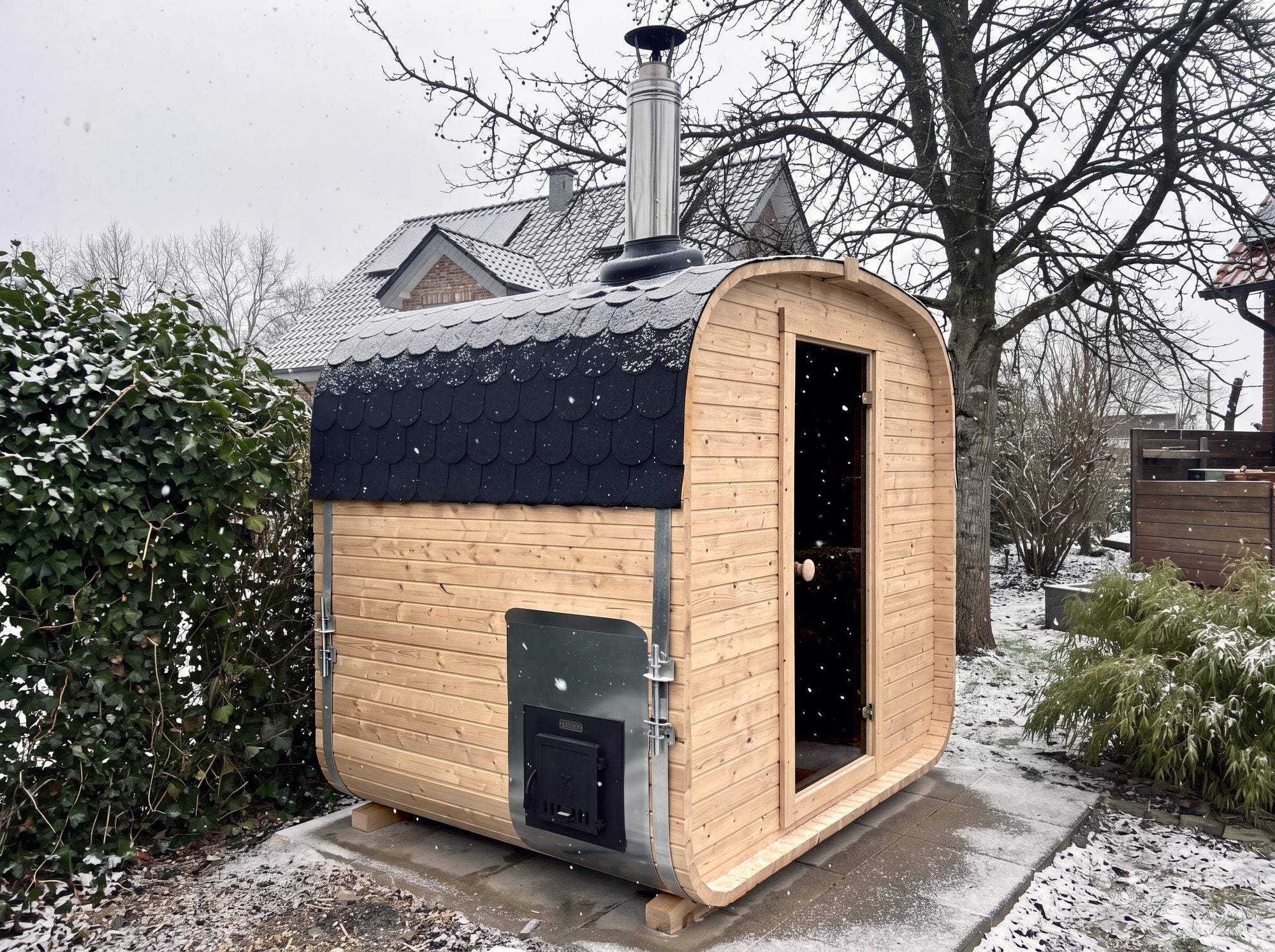 Wood-fired outdoor sauna with its flue and side firebox hatch in a snowy garden