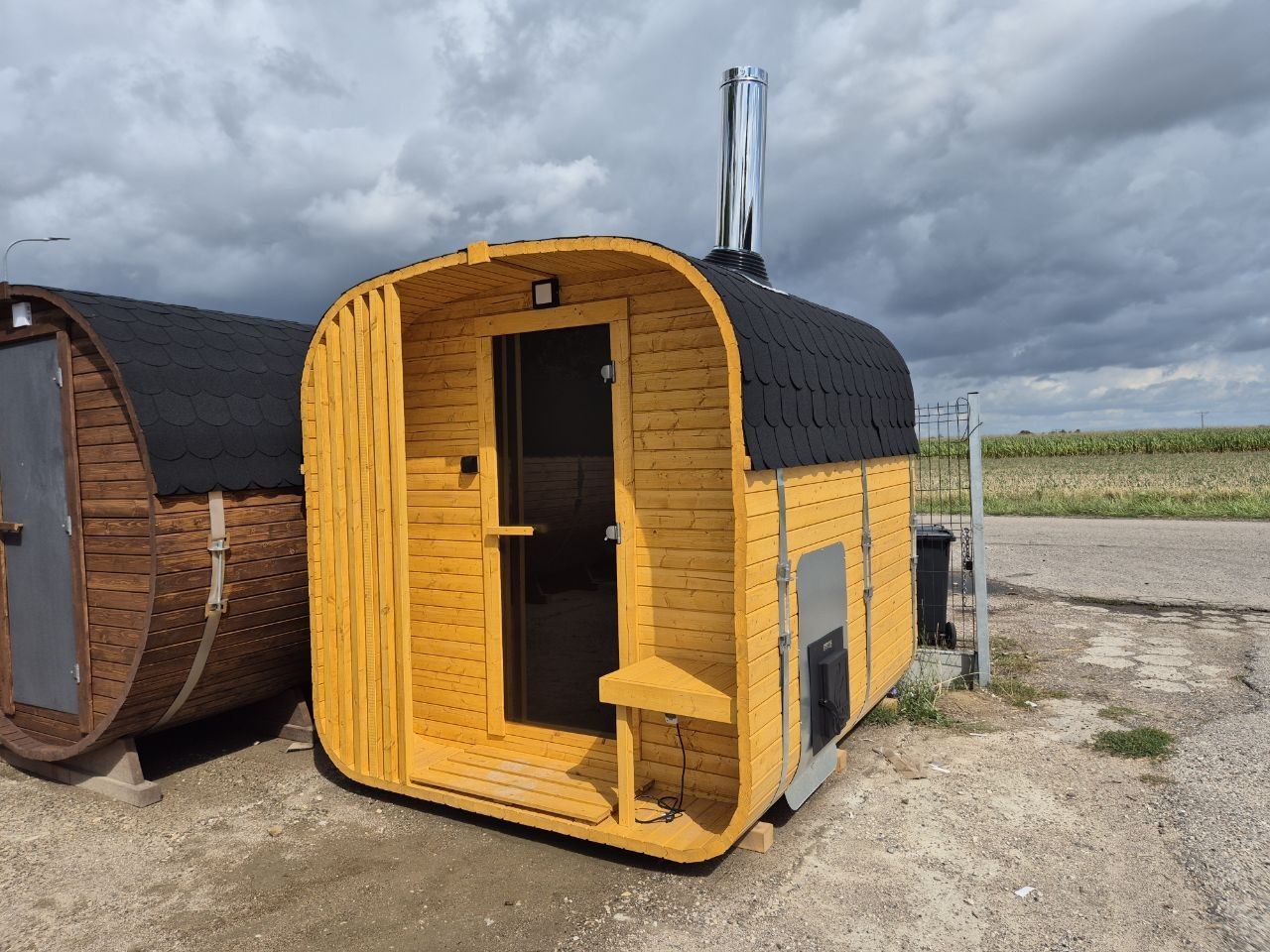 A barrel sauna and a cube-shaped outdoor sauna side by side, the cube with a stainless flue